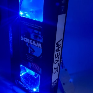 Może przedstawiać: Czarna kaseta VHS z filmem "Scream" i niebieskimi światłami LED. Taśma ma białą etykietę z napisem "Scream" w pionie. Światła oświetlają wnętrze i zewnętrzną stronę taśmy.