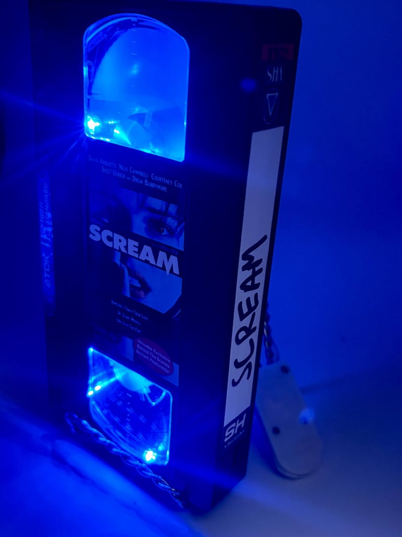 Może przedstawiać: Czarna kaseta VHS z filmem "Scream" i niebieskimi światłami LED. Taśma ma białą etykietę z napisem "Scream" w pionie. Światła oświetlają wnętrze i zewnętrzną stronę taśmy.