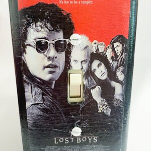 May include: A black light switch cover featuring the movie poster for "The Lost Boys." The poster art includes several people, with the text "Sleep all day. Party all night. Never grow old. Never die. It's fun to be a vampire."