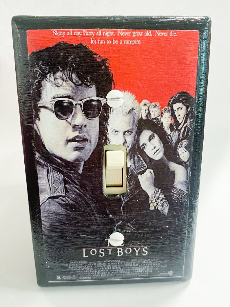 May include: A black light switch cover featuring the movie poster for "The Lost Boys." The poster art includes several people, with the text "Sleep all day. Party all night. Never grow old. Never die. It's fun to be a vampire."
