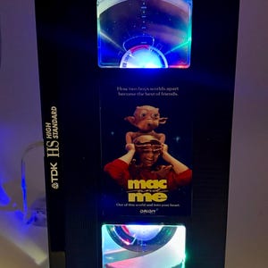May include: Black TDK HS High Standard VHS tape player with a "Mac and Me" movie poster. The player has three illuminated sections with blue, green, and red lights. The movie poster has the text "How two boys worlds apart become the best of friends."