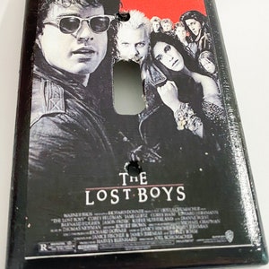 May include: Rectangular light switch cover featuring the movie poster for "The Lost Boys." The poster art includes images of several actors and the movie title. The top of the cover has the text: "Sleep all day. Party all night. Never grow old. Never die. It's fun to be a vampire."
