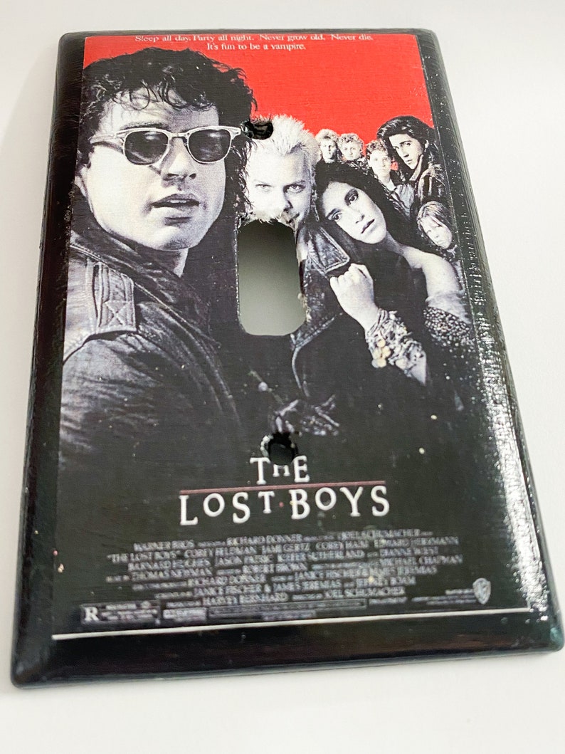 May include: Rectangular light switch cover featuring the movie poster for "The Lost Boys." The poster art includes images of several actors and the movie title. The top of the cover has the text: "Sleep all day. Party all night. Never grow old. Never die. It's fun to be a vampire."