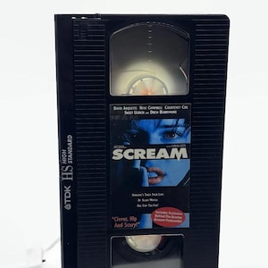 Może przedstawiać: Czarna kaseta VHS z filmem "Scream" firmy TDK. Na taśmie znajduje się tytuł filmu i nazwiska aktor&oacute;w na etykiecie. Etykieta zawiera r&oacute;wnież hasło "Clever, Hip And Scary!"