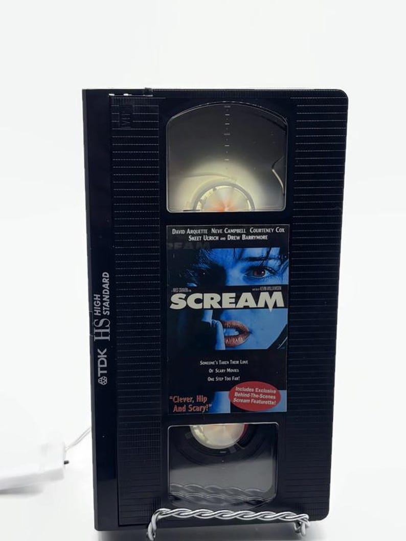 Może przedstawiać: Czarna kaseta VHS z filmem "Scream" firmy TDK. Na taśmie znajduje się tytuł filmu i nazwiska aktor&oacute;w na etykiecie. Etykieta zawiera r&oacute;wnież hasło "Clever, Hip And Scary!"