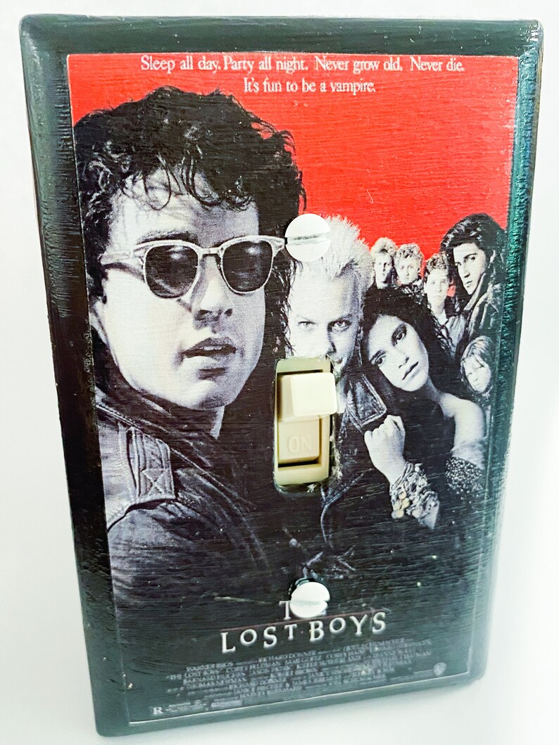 May include: A black light switch cover featuring the movie poster for "The Lost Boys." The poster art includes images of the film's characters, with the text "Sleep all day. Party all night. Never grow old. Never die. It's fun to be a vampire."