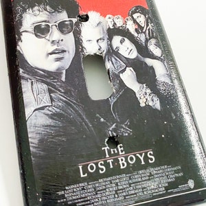 May include: Black light switch cover featuring the movie poster for "The Lost Boys." The poster art includes images of several actors and the movie title. The top of the cover has the text: "Sleep all day. Party all night. Never grow old. Never die. It's fun to be a vampire."