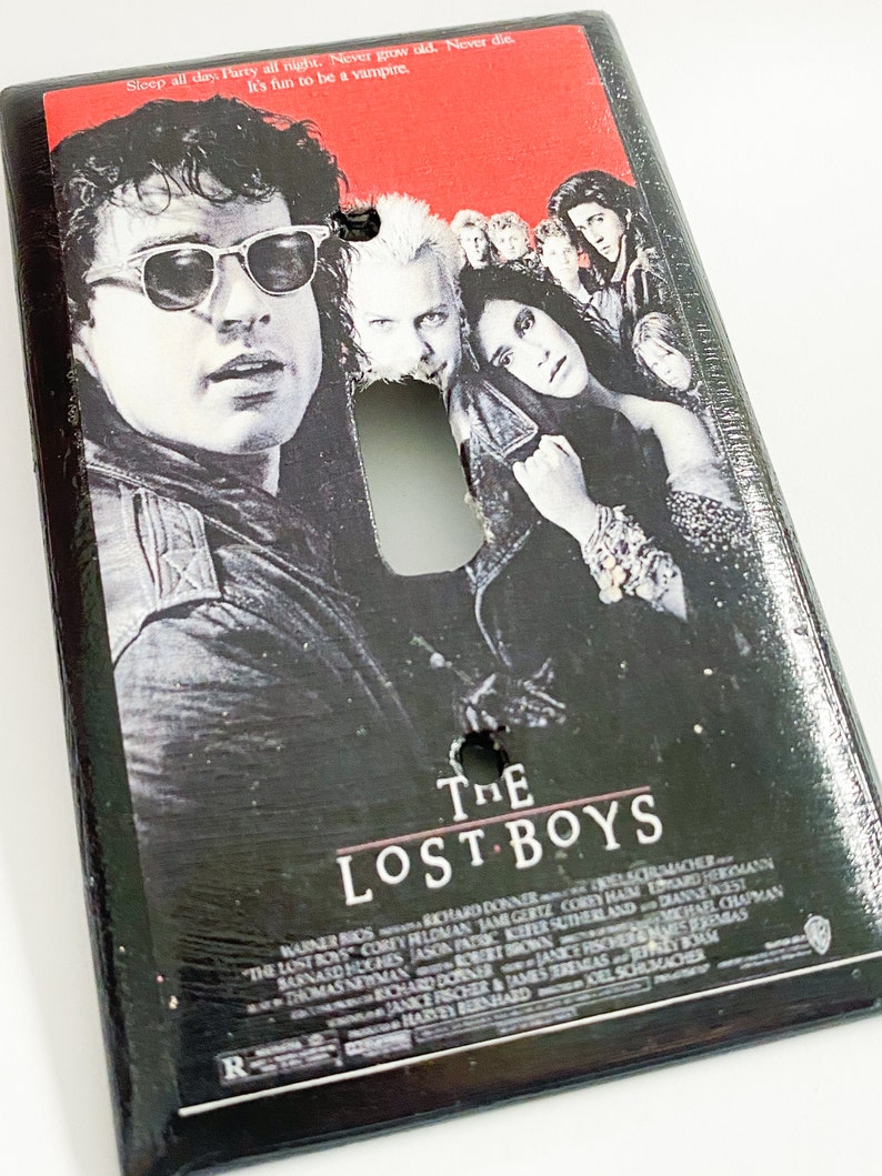 May include: Black light switch cover featuring the movie poster for "The Lost Boys." The poster art includes images of several actors and the movie title. The top of the cover has the text: "Sleep all day. Party all night. Never grow old. Never die. It's fun to be a vampire."