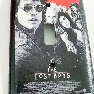 May include: A rectangular light switch cover featuring the movie poster for "The Lost Boys." The poster art includes images of several actors and the film's title in white lettering. The top of the cover has the text: "Sleep all day. Party all night. Never grow old. Never die. It's fun to be a vampire."