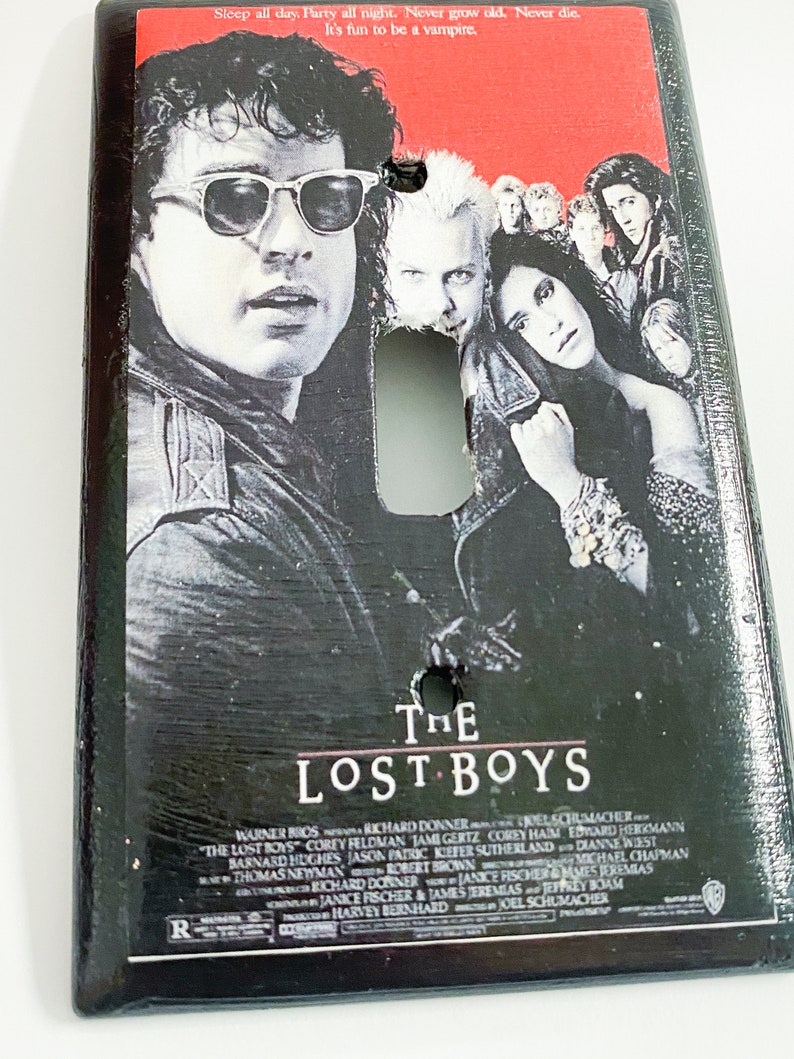 May include: A rectangular light switch cover featuring the movie poster for "The Lost Boys." The poster art includes images of several actors and the film's title in white lettering. The top of the cover has the text: "Sleep all day. Party all night. Never grow old. Never die. It's fun to be a vampire."