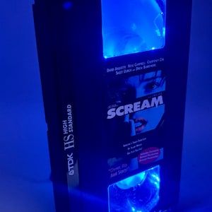 May include: A black VHS tape case with blue LED lighting. The case features the movie title "Scream" and the names of the actors. The tape is labeled "TDK HS HIGH STANDARD". The case has a clear, arched window.