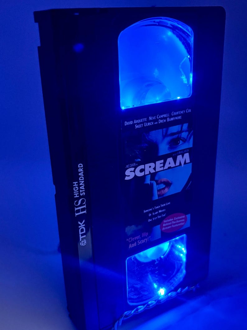 Może przedstawiać: Czarna obudowa kasety VHS z niebieskim oświetleniem LED. Obudowa zawiera film "Scream" i nazwiska aktor&oacute;w. Taśma jest oznaczona jako "TDK HS HIGH STANDARD". Posiada przezroczyste, łukowate okno.