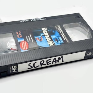 Może przedstawiać: Czarna kaseta VHS z napisem "Scream" napisanym czarnym markerem. Taśma ma przezroczyste okienko pokazujące szpule w środku. Okładka filmu przedstawia zbliżenie twarzy.