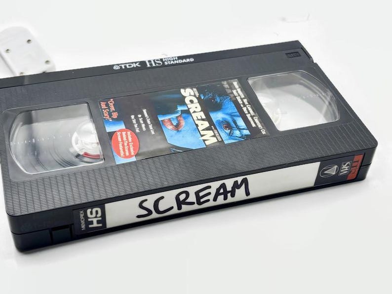 Może przedstawiać: Czarna kaseta VHS z napisem "Scream" napisanym czarnym markerem. Taśma ma przezroczyste okienko pokazujące szpule w środku. Okładka filmu przedstawia zbliżenie twarzy.