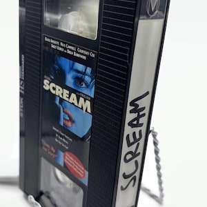 Może przedstawiać: Czarna kaseta VHS z filmem "Scream" z białym tytułem z boku. Taśma jest umieszczona na srebrnym drucianym stojaku. Okładka filmu przedstawia niebieski obraz twarzy.