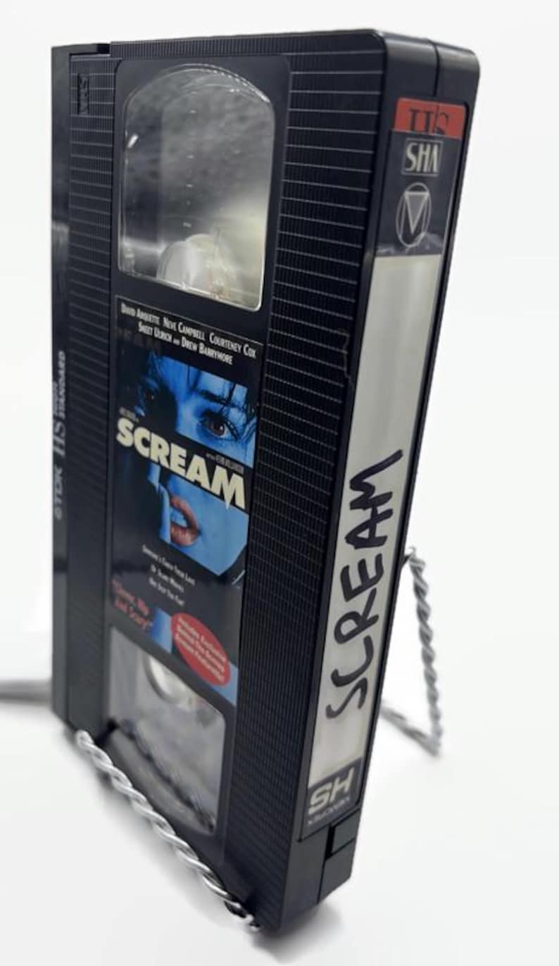 Może przedstawiać: Czarna kaseta VHS z filmem "Scream" z białym tytułem z boku. Taśma jest umieszczona na srebrnym drucianym stojaku. Okładka filmu przedstawia niebieski obraz twarzy.