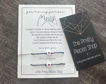 May include: Two matching friendship bracelets with a heart charm, displayed with a card that reads "you're my person" and a black tag with the shop's name. The bracelets have a black cord and a white heart with a red heart in the center.