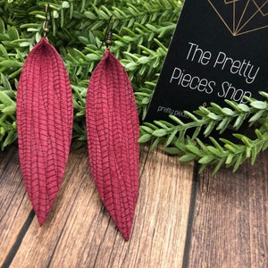 Peut inclure: Une paire de boucles d'oreilles en cuir bordeaux en forme de plumes. Les boucles d'oreilles ont un motif texturé et sont suspendues à des crochets dorés.