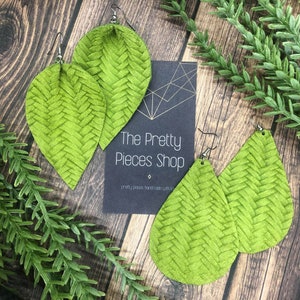 Op de afbeelding: Een paar groene leren oorbellen met een geweven patroon. De oorbellen zijn traanvormig en hebben zilveren haakjes. De oorbellen worden getoond op een houten oppervlak met een bord waarop "The Pretty Pieces Shop" staat.