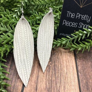 May include: A pair of light gray, leaf-shaped earrings with a textured chevron pattern. The earrings hang from silver-colored hooks. The background includes greenery and a wooden surface, with a black tag that reads "The Pretty Pieces Shop."