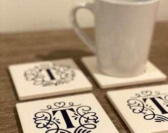 May include: Four white ceramic coasters with a black floral design and the letter "T" in the center. The coasters are on a wooden surface next to a white ceramic mug.