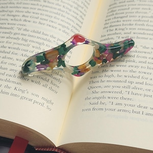 May include: A colorful, multi-faceted ring made of resin with embedded stones. The ring is pink, green, purple, and yellow. The ring is resting on an open book.