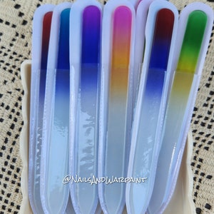 May include: Set of seven glass nail files with ombre colored tips. The colors are red, blue, purple, yellow, green, and two shades of blue. The files are in a white plastic container. The text "@NailsAndWarPaint" is visible on the container.