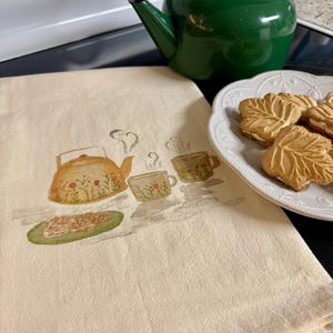 May include: A cream-colored tea towel with a watercolor illustration of a teapot, teacups, and a plate of food. A green teapot and a plate of leaf-shaped cookies are also visible.