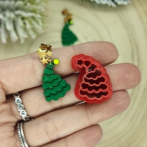 Könnte beinhalten: Grüne Weihnachtsbaum-Ohrringe mit goldenen Sternakzenten und eine rote Weihnachtsmützen-Brosche. Der Schmuck wird auf einer Holzoberfläche mit festlichem Grün im Hintergrund präsentiert.