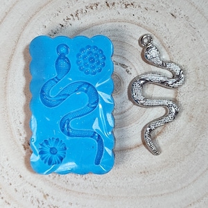 Puede incluir: Un molde de silicona azul con un diseño de serpiente y dos formas de flores. También se muestra un amuleto de serpiente plateado.