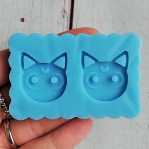 May include: Blue silicone mold with two cat face cavities. Each cavity has a crescent moon on the forehead and two round eyes.