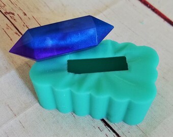 Puede incluir: Un molde de silicona verde azulado en forma de nube con una abertura rectangular. Un cristal azul y morado está sentado encima del molde.