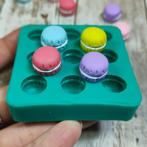Può includere: Uno stampo in silicone turchese pieno di macaron in miniatura colorati. I macaron sono nelle tonalità di blu, rosa, giallo e viola, con ripieno bianco. Diversi macaron sciolti sono sparsi intorno allo stampo. L'immagine è ripresa da vicino.