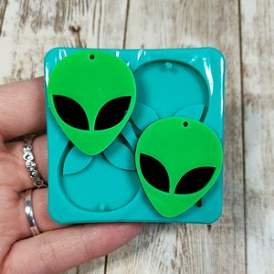 May include: A green silicone mold for making two alien-shaped earrings. The mold is turquoise blue and has two cavities shaped like aliens with black eyes.