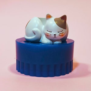 Può includere: Un temperamatite di plastica blu con una statuetta di gatto bianco e marrone che dorme in cima. Il gatto ha gli occhi chiusi e un naso rosa.