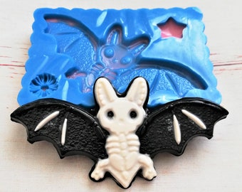 Puede incluir: Un murciélago de resina blanca con alas negras y un diseño esquelético. El murciélago está sentado sobre un molde de silicona azul con una estrella y un diseño de murciélago.