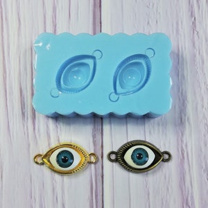 Puede incluir: Dos amuletos de ojo turco azul y blanco con engastes dorados y bronce. Los amuletos se muestran junto a un molde de silicona con dos formas de ojo.
