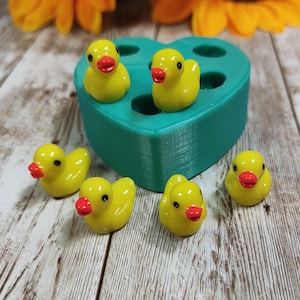 May include: A teal heart-shaped silicone mould with multiple circular cavities, accompanied by five small yellow rubber duck figurines featuring orange beaks and black eyes, arranged around the base of the mould on a light wood surface.