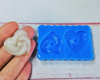 May include: A close-up of a blue silicone mould with two heart-shaped cavities. The mould is used to create resin hearts with a swirl design. A white resin heart with a swirl design is shown next to the mould.