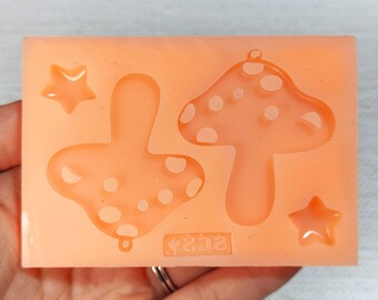 May include: A silicone mold for making resin crafts. The mold features two mushroom shapes with white polka dots and two star shapes. The mold is light orange in color and has the text "3233" printed on the bottom.