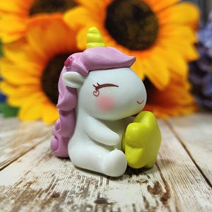 Puede incluir: Una pequeña figura de unicornio blanco con pelo rosa y una estrella amarilla. El unicornio tiene los ojos cerrados y un rubor rosa en las mejillas.