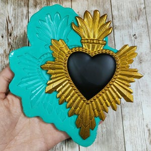 Peut inclure: Un emblème religieux doré en forme de cœur avec un centre noir, entouré de rayons et d'une couronne. L'emblème est posé sur un moule en silicone de couleur turquoise avec un motif floral. Le fond est une surface en bois.