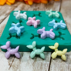 Puede incluir: Un molde de silicona verde con nueve cavidades con forma de perros globo. El molde está lleno de figuritas de perros globo de resina de color pastel. Las figuritas son de varios colores, incluyendo rosa, azul, amarillo y verde.