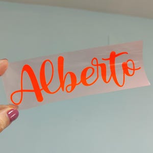Può includere: Un foglio di trasferimento adesivo trasparente rettangolare con il nome Alberto scritto in un carattere corsivo arancione brillante. Le lettere sono audaci e fluide, progettate per essere applicate su una superficie liscia come adesivo.