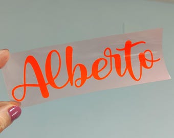 May include: A rectangular piece of clear transfer tape featuring the name Alberto written in a vibrant orange, cursive script font. The lettering is bold and fluid, with the capital A and lowercase letters connected in a single, continuous style.