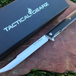 May include: A black folding knife with a silver blade and a black handle. The knife is open and the blade is visible. The knife is on a brown surface with green grass in the background. The knife has the text "Tactical Gearz" on the blade and the box.