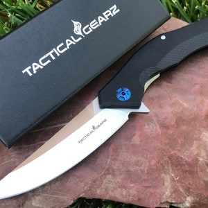 May include: A black pocket knife with a silver blade and a blue accent. The knife is open and the blade is sharp. The knife is on a brown rock with green grass in the background. The knife has the text "Tactical Gearz" on the blade and the box.