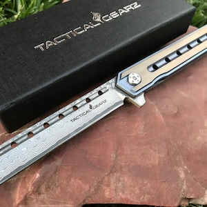 May include: A silver and gold tactical knife with a Damascus steel blade and a black box with the text "Tactical Gearz" on it.