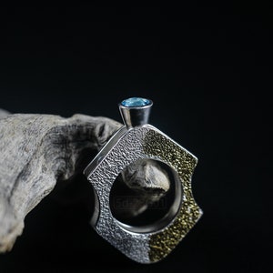 Contemporary ring with reticulated sterling silver and keum boo 24k gold foil and blue topaz stone, double-sided. On Black Background, Next to a Piece of Dry Cactus Wood.