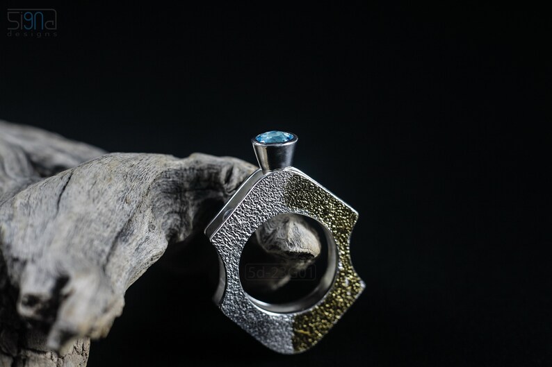 Contemporary ring with reticulated sterling silver and keum boo 24k gold foil and blue topaz stone, double-sided. On Black Background, Next to a Piece of Dry Cactus Wood.